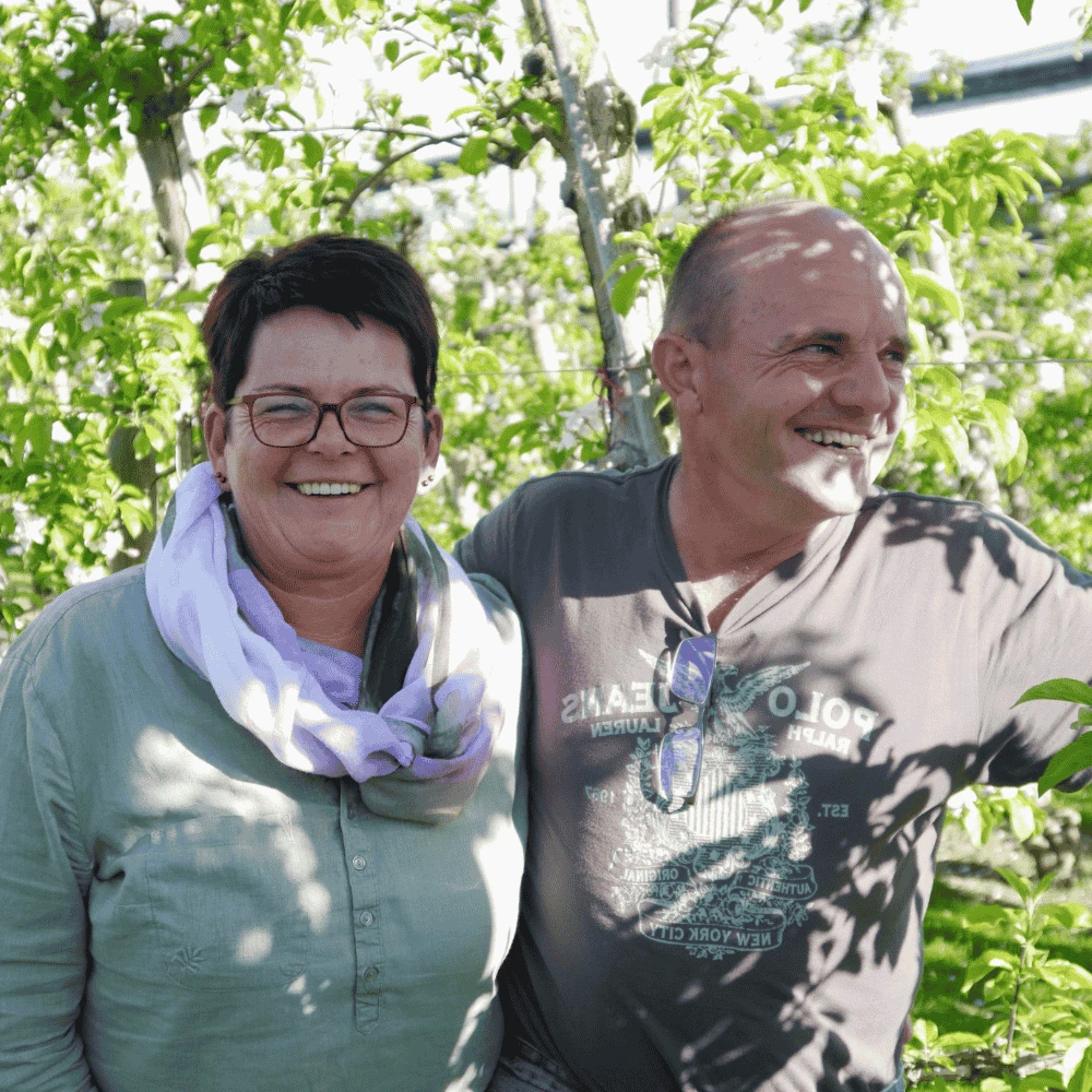 Die Gastgeber des Obstgut Ledinegg stehen lachend in ihrem blühenden Obstgarten in der Südsteiermark.