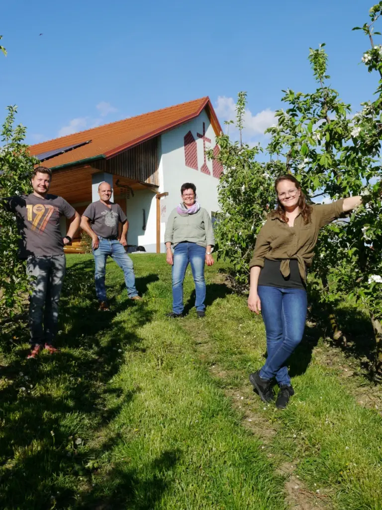 In der Obstanlage vom Obstgut Ledinegg posiert die Familie Ledinegg. Die zwei Eltern stehen in der Mitte. Die Tochter steht auf der rechten Seite und lehnt sich an einen Apfelbaum an. Der Schwiegersohn befindet sich auf der linken Seite und steht im Schatten der linken Apfelreihe.