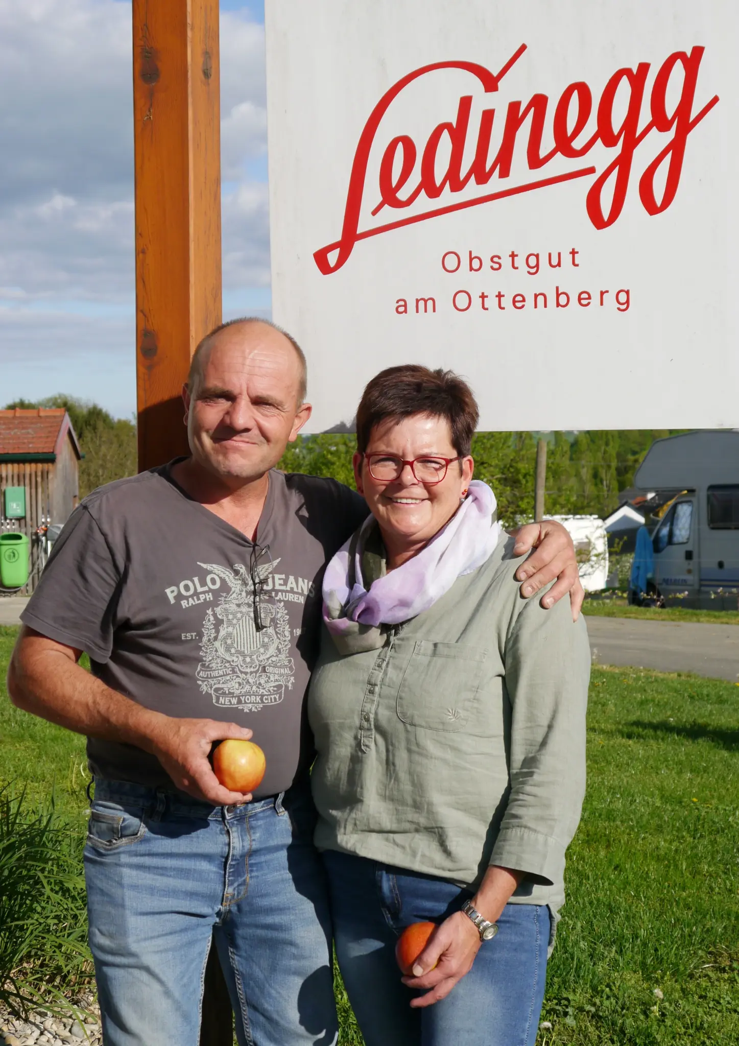 Das Gastgeber-Ehepaar Ledinegg steht lächelnd vor dem großen Hofschild "Ledinegg - Obstgut am Ottenberg", mit frischen Äpfeln in der Hand und Campingwagen im Hintergrund